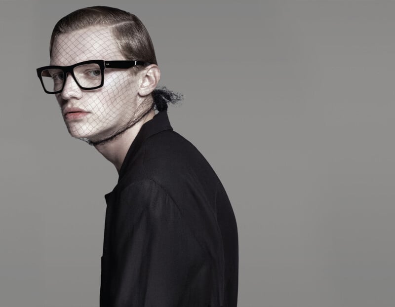 A person with slicked-back hair and large black glasses wears a black jacket and a mesh veil over their face and neck, set against a plain gray background. They look slightly to the side with a neutral expression.