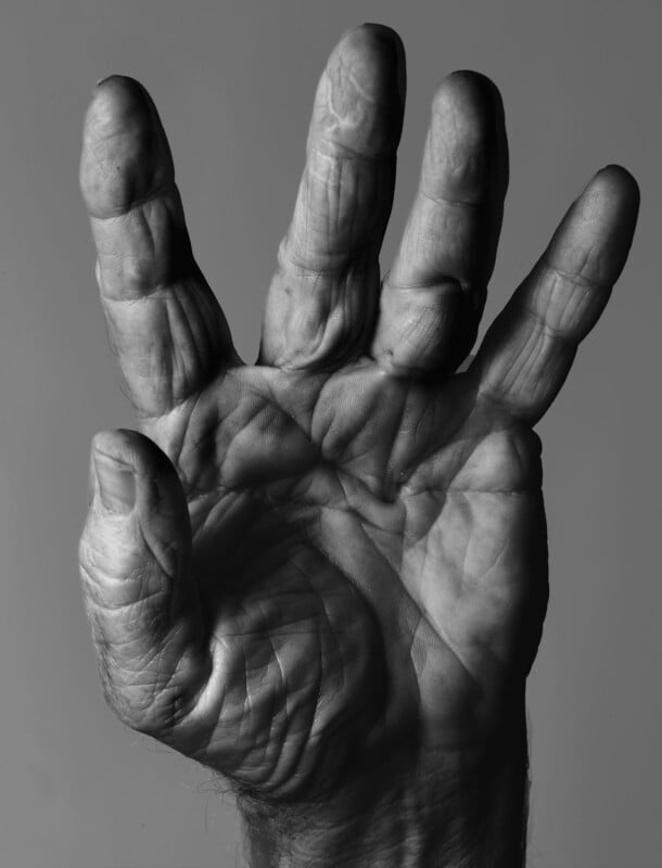 A close-up, black and white photo of an open hand with fingers spread apart, showing detailed lines and wrinkles on the palm and fingers against a plain background.