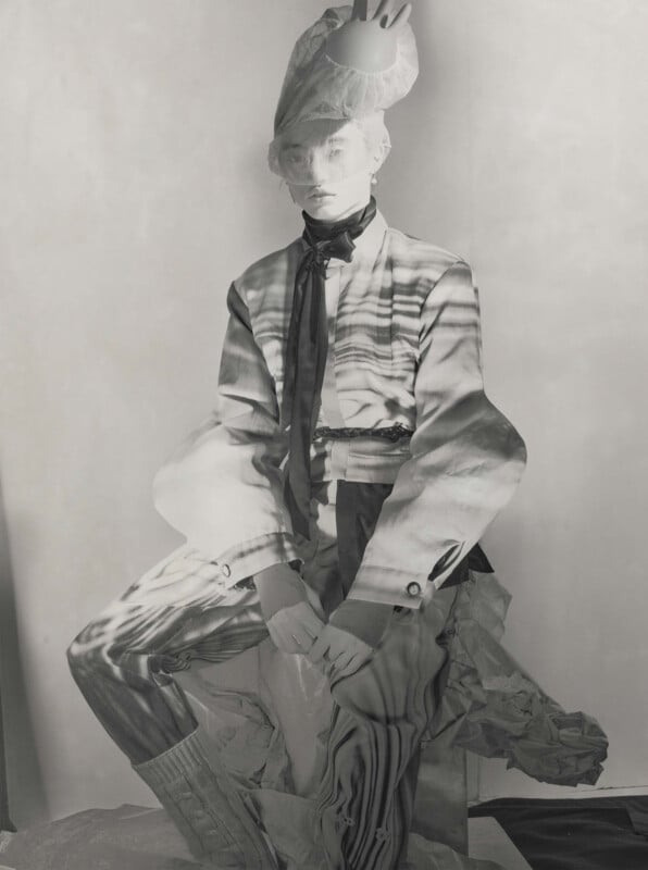 A person poses seated, wearing a surreal outfit with a patterned blazer, scarf, and sculptural headpiece shaped like a hand. Black and white lighting creates dramatic shadows and abstract patterns across their body and face.