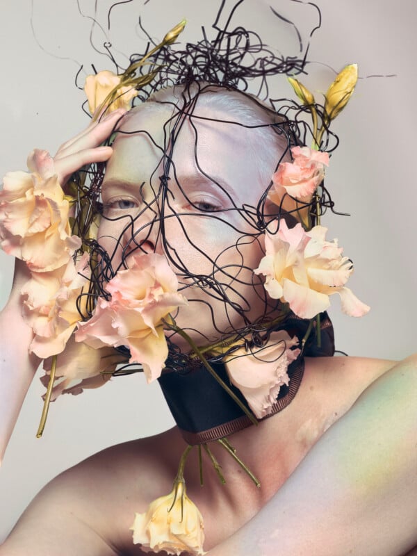 A person with pale painted skin wears a sculptural headpiece made of tangled black wire and pale pink flowers, including around their neck and hand, creating an avant-garde, ethereal look.