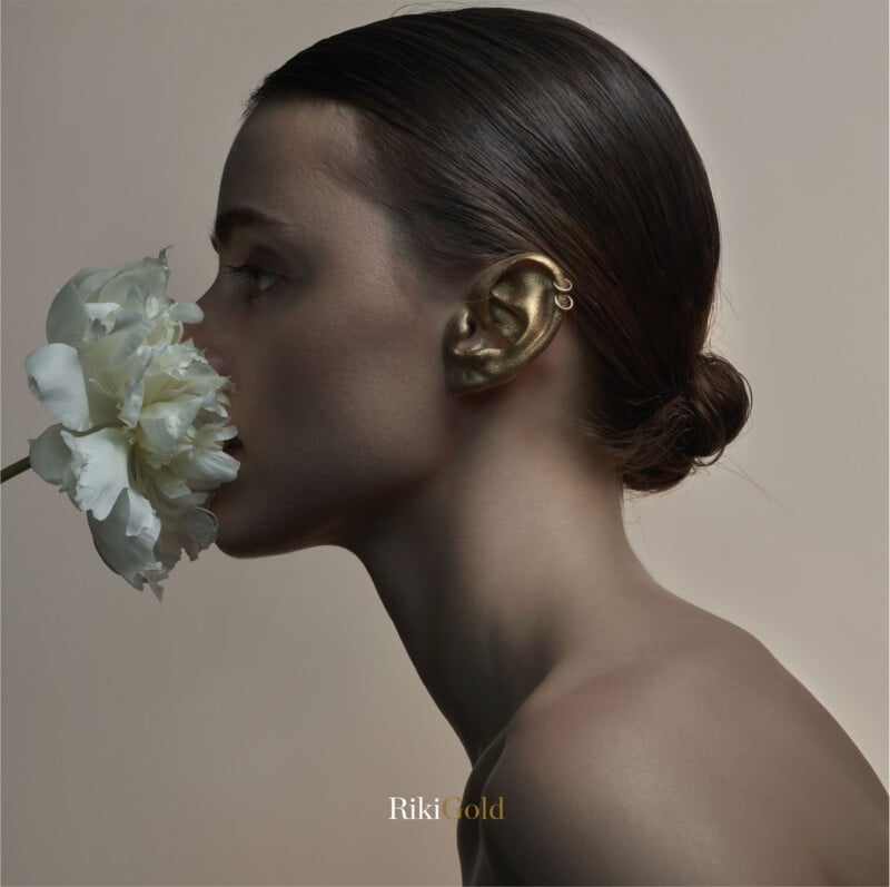 A woman with slicked-back hair holds a white flower to her face. Her ear is painted gold and adorned with two gold hoops. The background is neutral, and "RikiGold" is written at the bottom of the image.