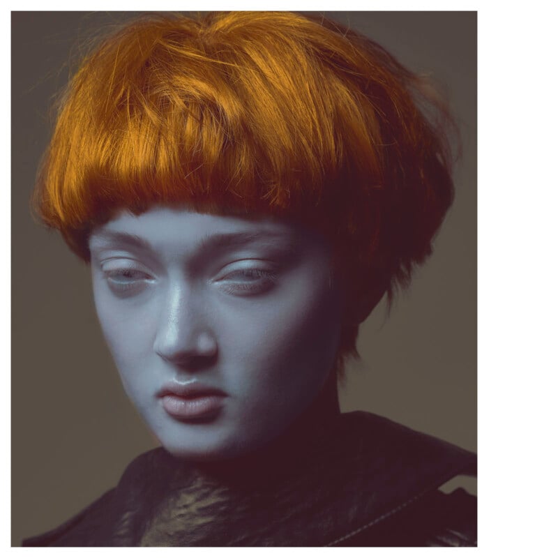 A person with short, bright orange hair and blue-painted skin looks downward with a neutral expression, wearing a dark-colored top against a plain background.