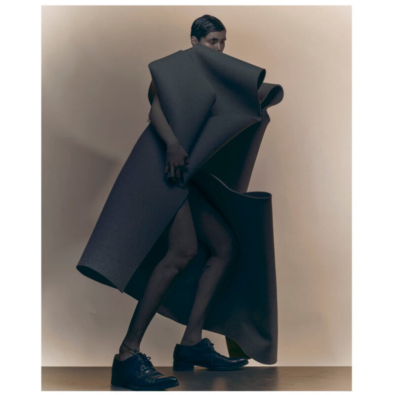 A person stands against a neutral background, wrapped in an oversized, sculptural piece of dark, textured fabric and wearing black dress shoes. Only their face, hands, and legs are visible.