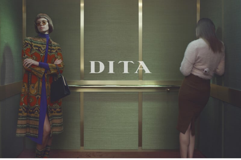 Two women stand in an elevator with green walls; one faces forward in a colorful coat and sunglasses, holding a bag, while the other faces the wall wearing a fuzzy sweater and brown skirt. The word "DITA" appears in the center.