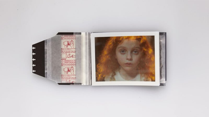 An open instant photo pack reveals a photograph of a young child with curly red hair and a solemn expression. The pack has instructions and red diagrams on the inner flap, all placed against a plain white background.