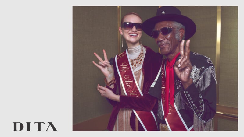 Two people wearing sunglasses and maroon sashes smile and flash peace signs. One wears a shiny outfit and cowboy hat; the other wears a velvet jacket. The DITA logo appears in the lower left corner.
