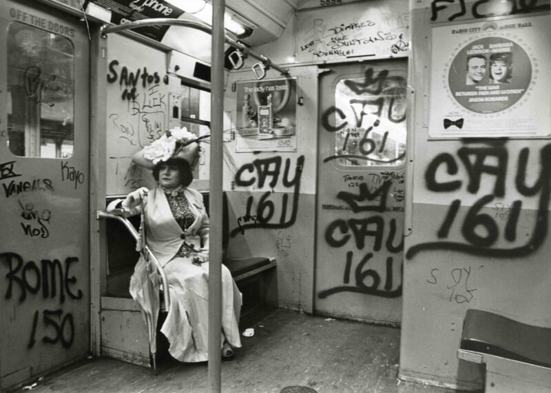 A person in a white dress and ornate hat sits alone in a subway car covered with graffiti, with posters and scribbles on the walls and doors.