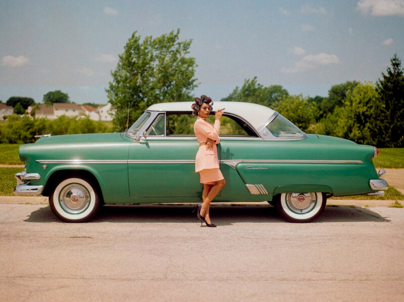 A woman in vintage attire stands beside a classic green and white car parked on a sunny street, with trees and houses in the background.