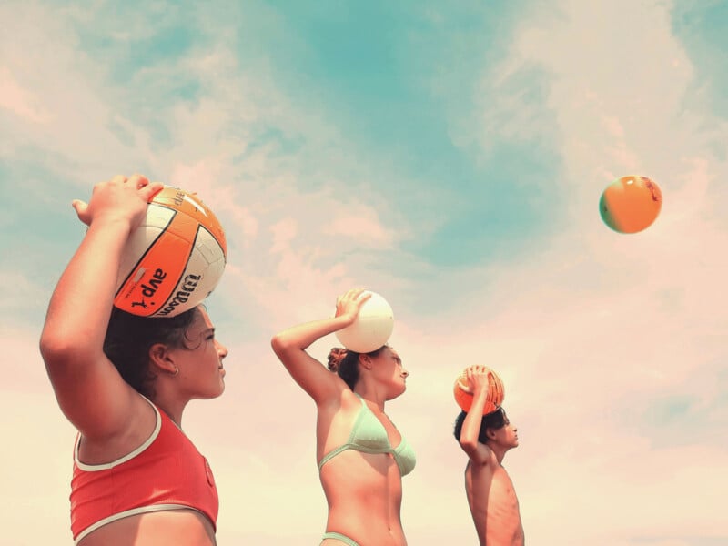 Three children in swimsuits hold volleyballs above their heads, preparing to throw, against a pastel sky with scattered clouds. The scene suggests a playful, sunny beach or outdoor setting.