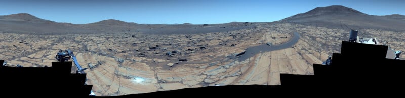 Panoramic view of Mars’ rocky, cracked surface with scattered boulders, hills in the distance, and part of a rover visible at the edge of the image under a clear blue sky.