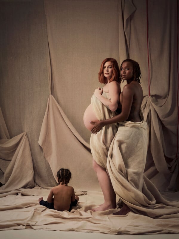 Two women stand wrapped in beige fabric, one visibly pregnant, while a small child with braided hair sits on the floor facing away. The background features draped beige cloth.