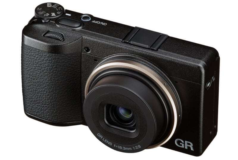 A black digital compact camera with a textured grip, control dial on top, and a protruding lens labeled "GR LENS f=18.3mm 1:2.8," facing slightly to the right on a white background.
