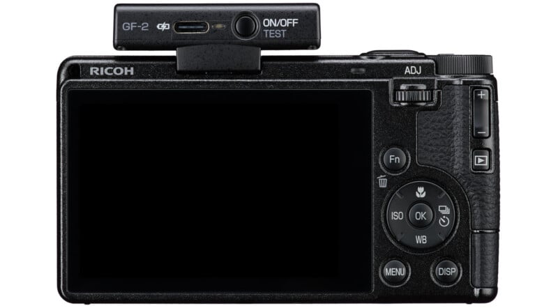 Rear view of a Ricoh digital camera with a large display screen, control buttons on the right side, and an attached rectangular accessory labeled "GF-2" on top with an ON/OFF TEST switch and USB-C port.