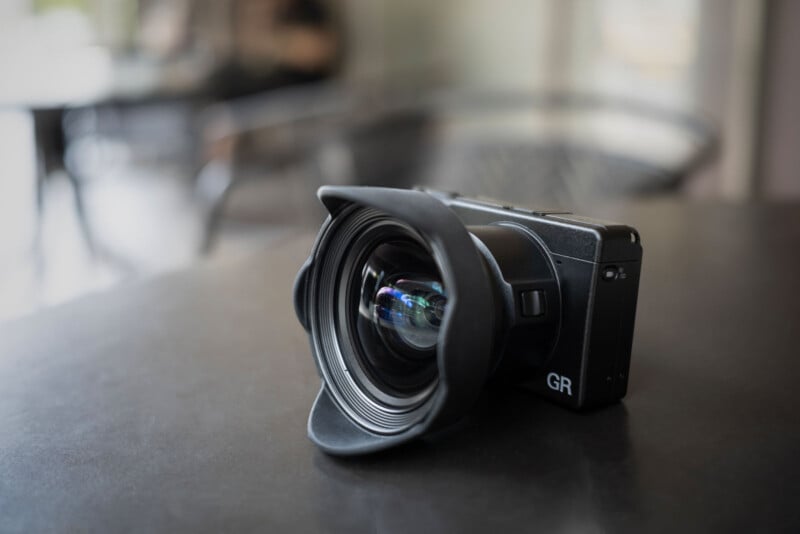 A black digital camera with a wide-angle lens attachment sits on a dark table, with a blurred background of chairs and a person.