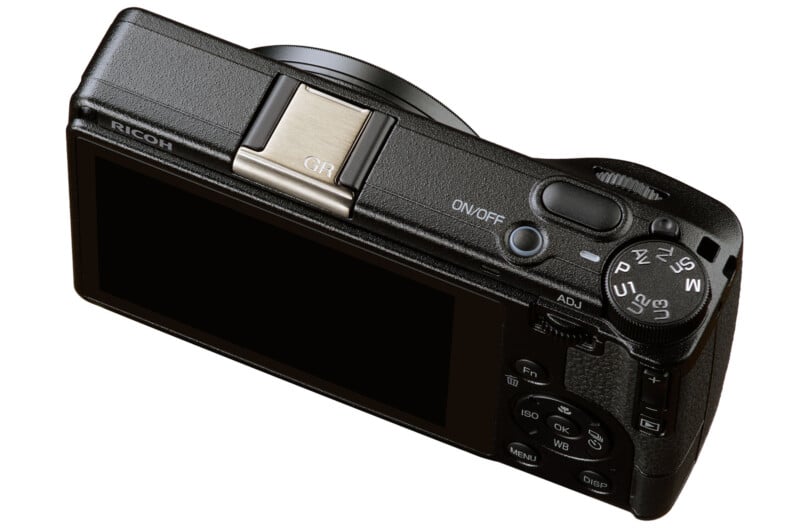 A black Ricoh digital camera viewed from the back, showing its LCD screen, control dial, and various buttons. The camera is angled slightly, revealing the lens barrel on the right.