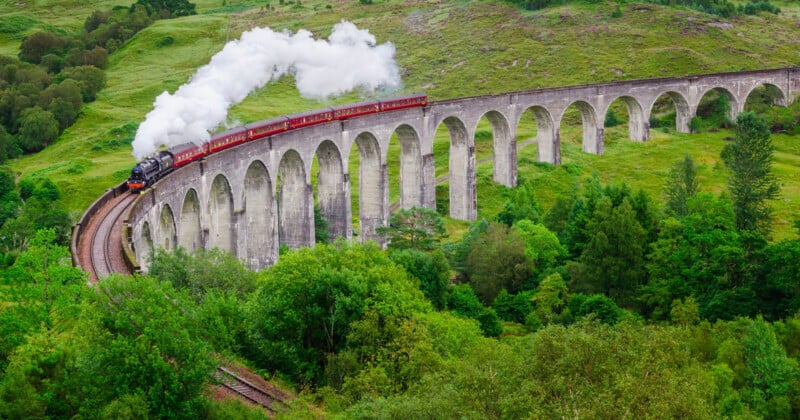 A steam train with red carriages crosses a tall, curved stone viaduct surrounded by lush green trees and rolling hills, with white smoke billowing from the engine.