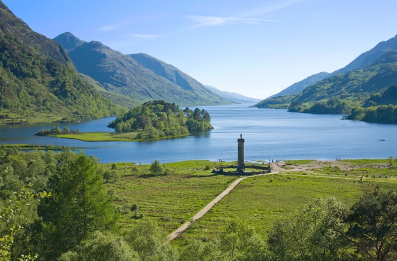 A scenic view of a monument by a lake, surrounded by green hills and mountains under a clear blue sky, with an island and lush vegetation in the background.