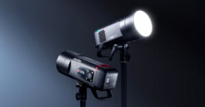Two professional studio lights on stands, one facing forward with its bright LED illuminated and the other showing its digital display screen and control buttons, both set against a dark gradient background.