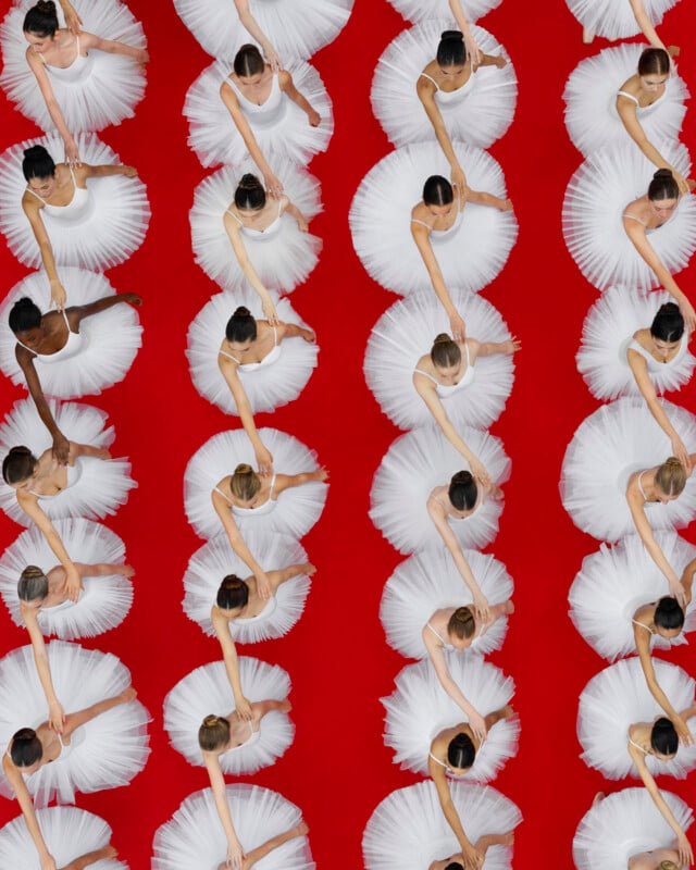 Overhead view of rows of ballerinas in white tutus, standing on a bright red floor, evenly spaced and posed with one arm curved gracefully above their heads.