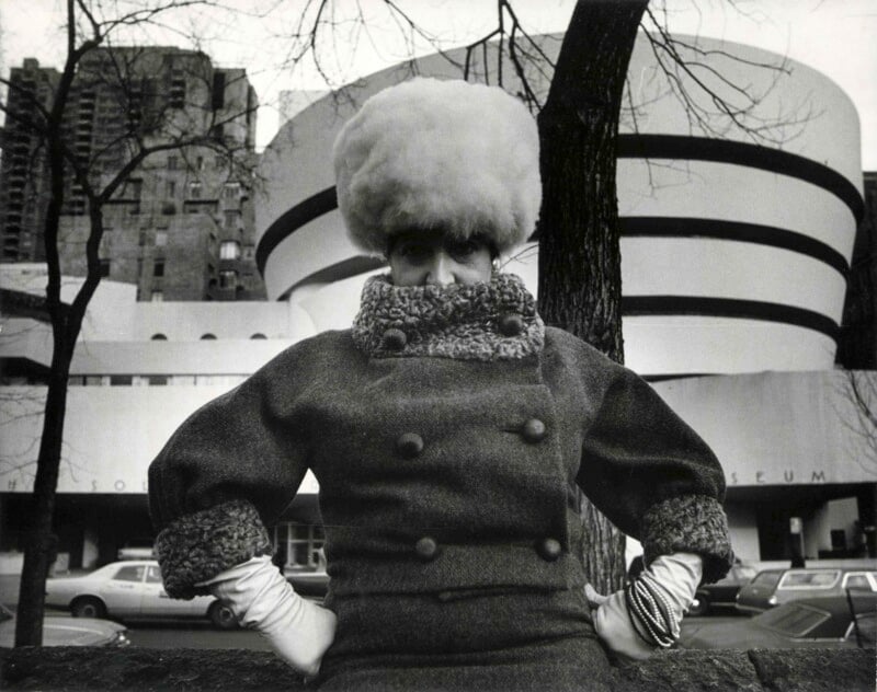 A person wearing a large fur hat, a heavy coat with a high collar, and white gloves poses in front of the Guggenheim Museum in New York City on a winter day, with bare trees and parked cars visible.