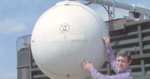 A man in a purple shirt stands next to a large, spherical white tank with a "W" logo on it, mounted on industrial equipment outdoors under a blue sky.