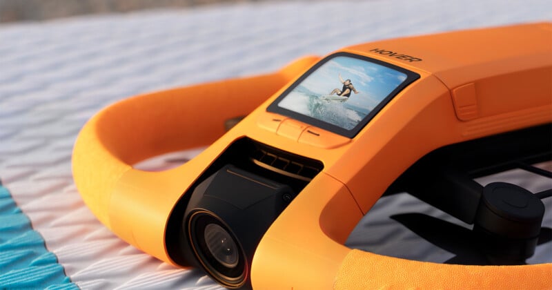 An orange underwater drone with a screen displaying a person surfing, placed on a textured white and blue surface.