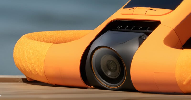 Close-up of an orange underwater drone with a camera lens, resting on a wooden surface, with a blurred blue background.