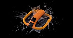 An orange underwater drone splashes through water against a black background, with droplets frozen in midair around its curved frame and camera lens.