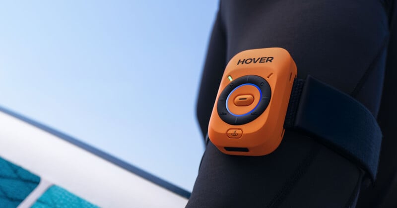 Close-up of an orange HOVER device with buttons, strapped to a person's black wetsuit-covered arm, with a blue sky and part of a pool or board visible in the background.
