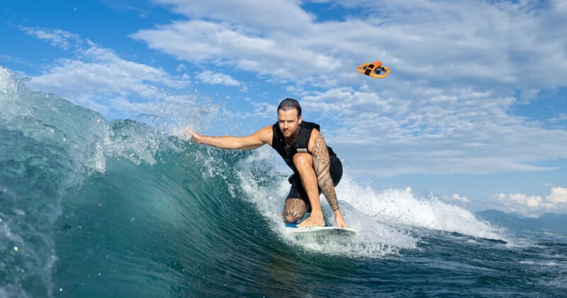 A man with tattoos surfs on a blue ocean wave under a partly cloudy sky, while an orange sandal flies through the air in the background.
