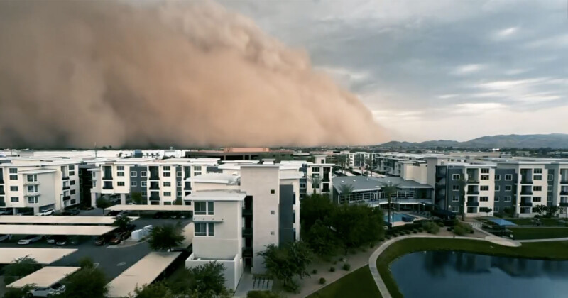 A massive dust storm, or haboob, approaches a modern residential complex with white and gray buildings, trees, and a small pond under a cloudy sky.