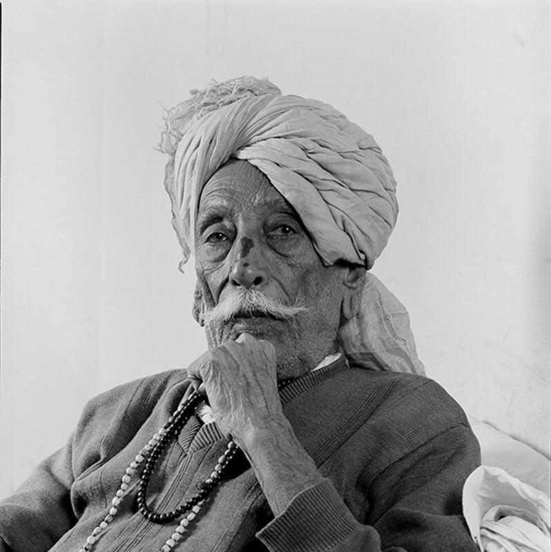 Black-and-white portrait of an elderly man with a mustache, wearing a traditional turban, a sweater, and beaded necklaces, sitting thoughtfully with his hand resting under his chin.
