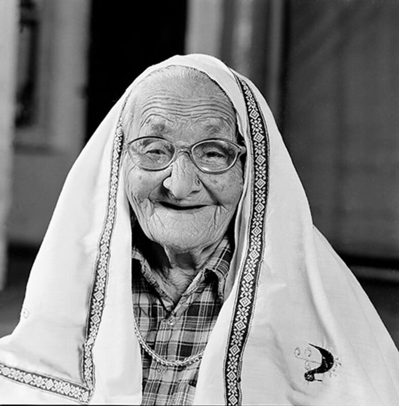 An elderly woman with glasses smiles warmly, wearing a patterned headscarf draped over her head and shoulders, and a checked blouse. The black-and-white photo highlights her gentle expression and the texture of her clothing.