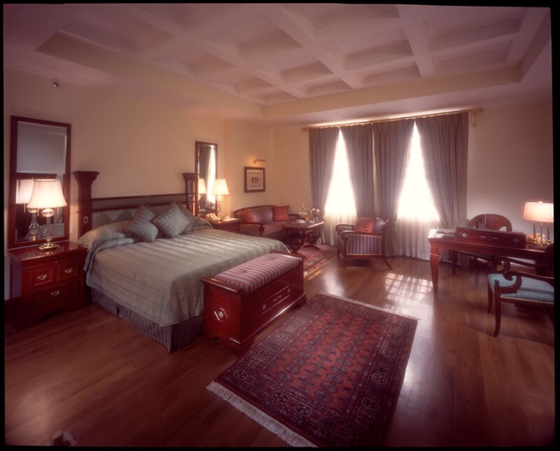 A spacious, elegant bedroom with a large bed, wooden furniture, a patterned rug, armchairs, a desk, and large windows with flowing curtains letting in natural light.