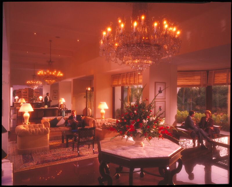 Luxurious hotel lobby with elegant chandeliers, plush sofas, lamps, a large floral arrangement on a marble table, and people relaxing or conversing. Warm lighting creates a cozy, inviting atmosphere with garden views through large windows.