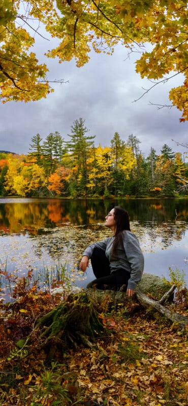 A person in a gray sweater sits on a rock by a calm lake, surrounded by colorful autumn trees with orange, yellow, and green leaves under a cloudy sky. Fallen leaves cover the ground and the lake reflects the vibrant foliage.