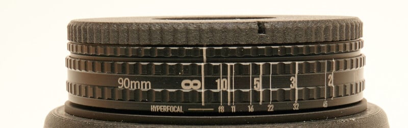 Close-up of the focus and aperture rings on a camera lens, showing measurement markings with numbers including 90mm, 8, 10, 5, and the word "HYPERFOCAL." The lens surface is textured and black.