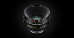 A professional camera lens with ridged focus and aperture rings, marked with white and green numbers, is displayed against a dark background. The lens features a red logo on its barrel.