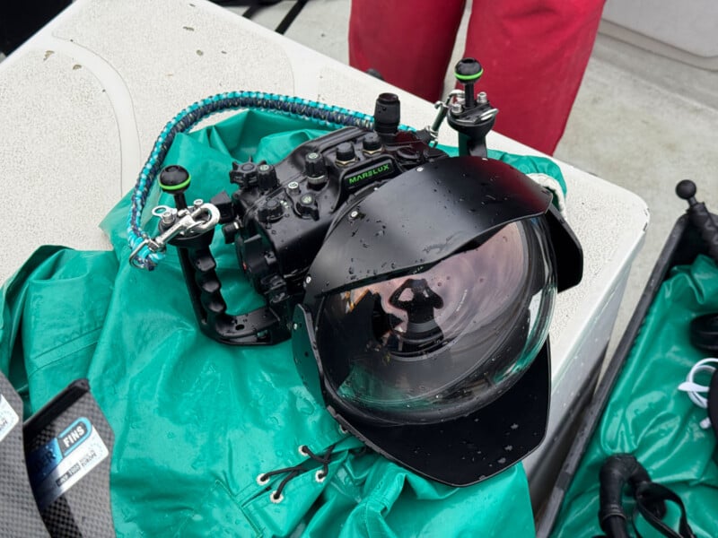 A professional underwater camera housing with attached controls and dome port sits on a green tarp, with droplets of water on it and a braided strap; a person's red pants are visible in the background.