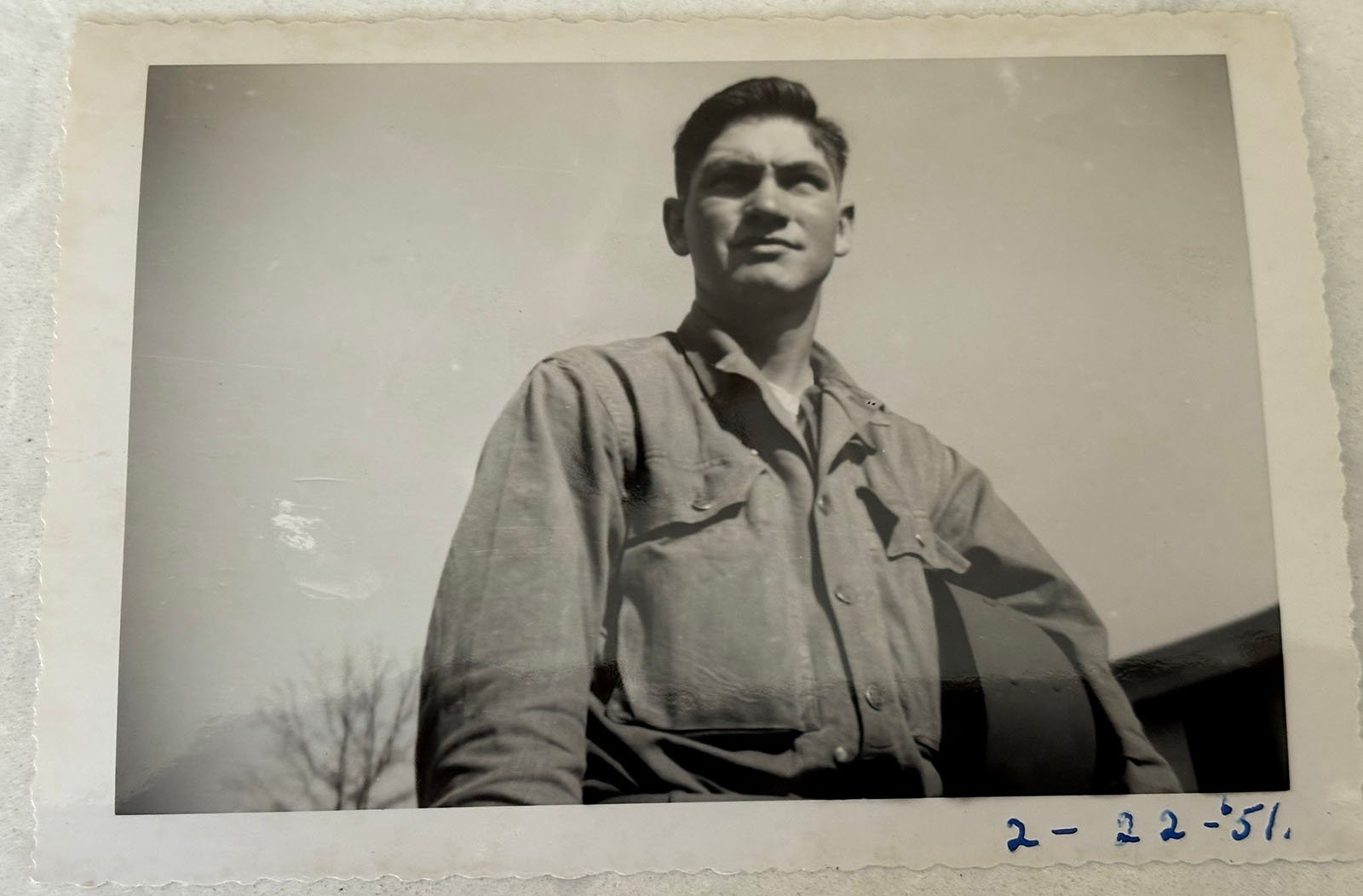 Black and white photo of a man in a button-up shirt holding a hat. He is looking slightly upward, posing outdoors. The date "2-22-51" is handwritten in blue ink on the photo’s white border.