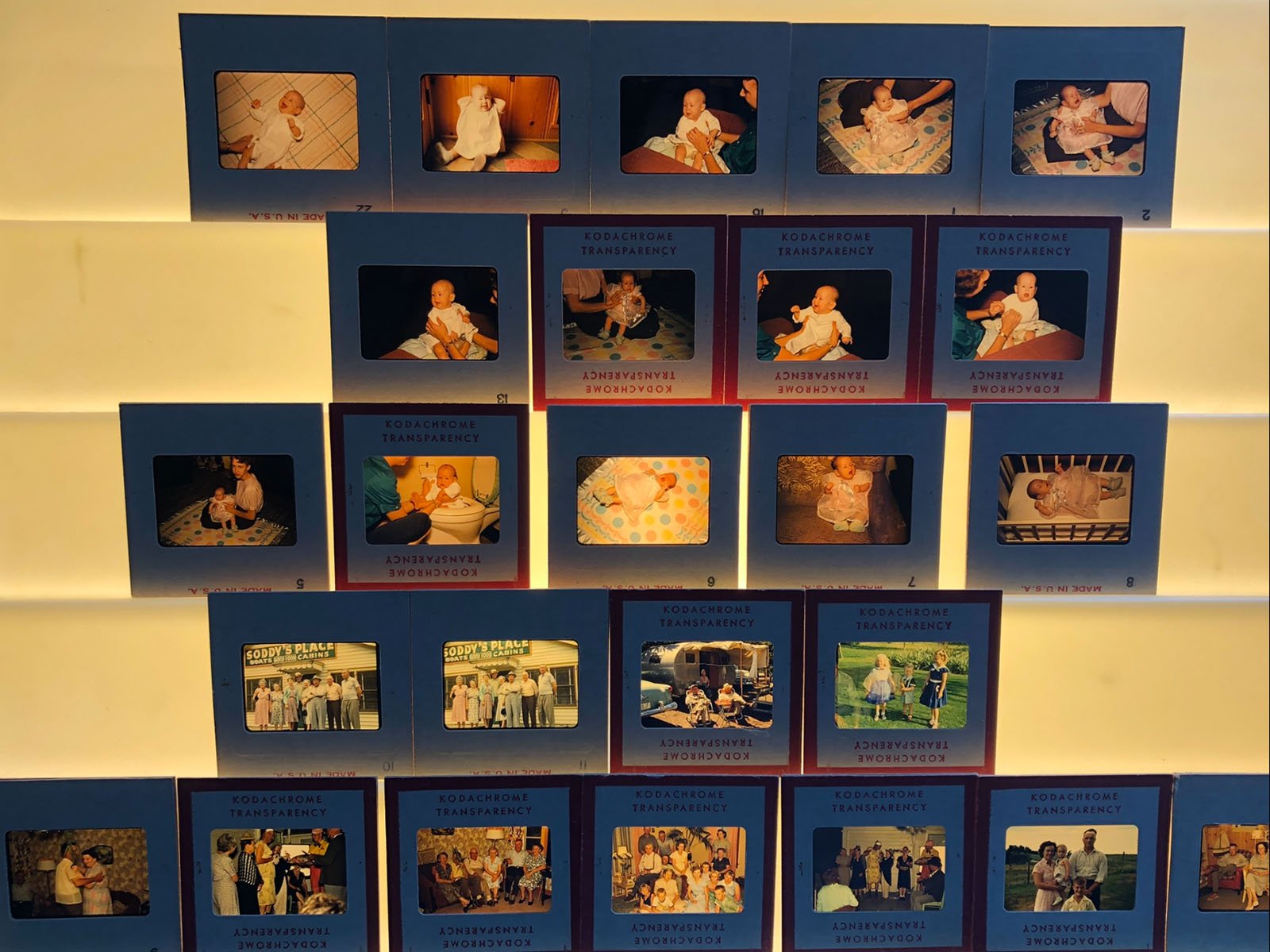 A collection of vintage Kodak slide transparencies displayed on a lightbox, showing various family moments including babies, toddlers, and adults indoors and outdoors. Each slide is labeled and arranged in rows.