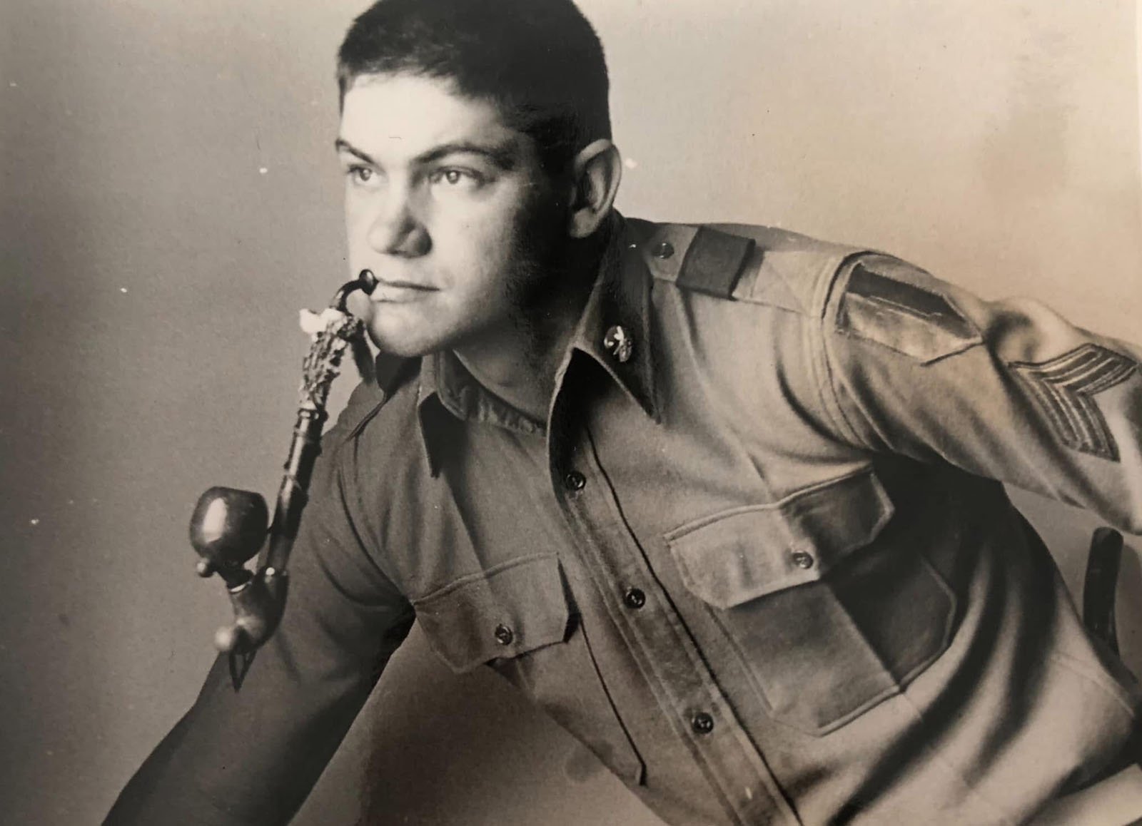 A man in a military uniform leans forward while playing a saxophone. The photo is black and white, and he appears focused, looking slightly to the side.