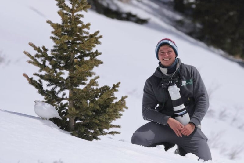 A person kneels in the snow beside a small evergreen tree, smiling at the camera. A white bird stands nearby. The person wears winter clothing and has a large camera with a telephoto lens. Snow and trees fill the background.
