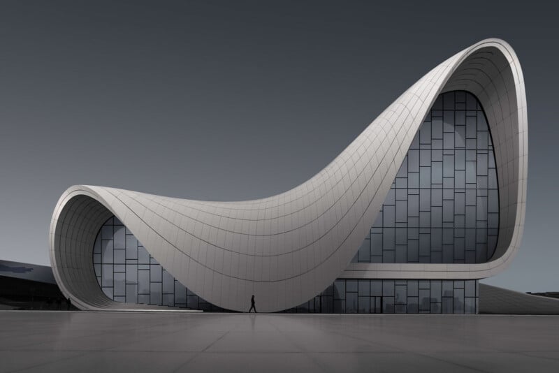 A modern building with smooth, curving lines and a wave-like roof, featuring large glass windows. A solitary person walks in front, highlighting the structure’s impressive, futuristic design against a gray sky.