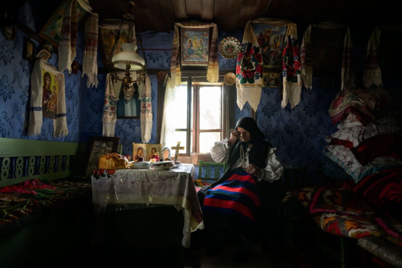 An elderly woman in traditional clothing sits near a window in a dimly lit, ornately decorated room with religious icons, embroidered textiles, and a table set with food and candles.