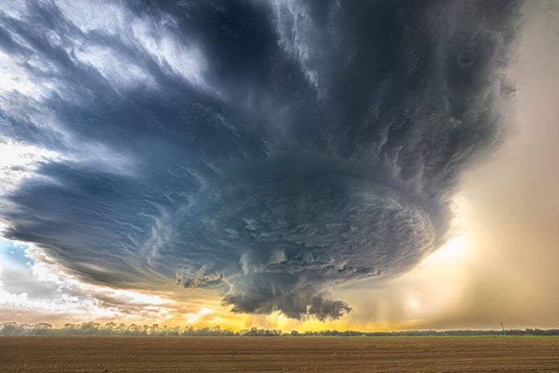 A dramatic supercell thunderstorm with swirling dark clouds hovers over a flat, empty field at sunset, with golden light illuminating the horizon.