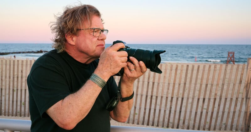 A person with glasses and tousled hair holds a camera, preparing to take a photo near a beach at sunset. A wooden fence and the ocean are visible in the background.
