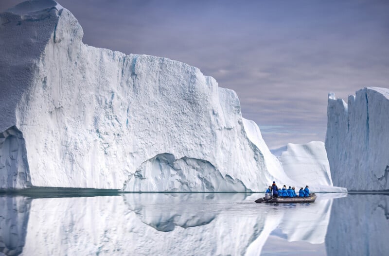 A small boat with people in blue jackets floats on calm water surrounded by large, towering white icebergs, with their reflections visible on the water’s surface under a cloudy sky.