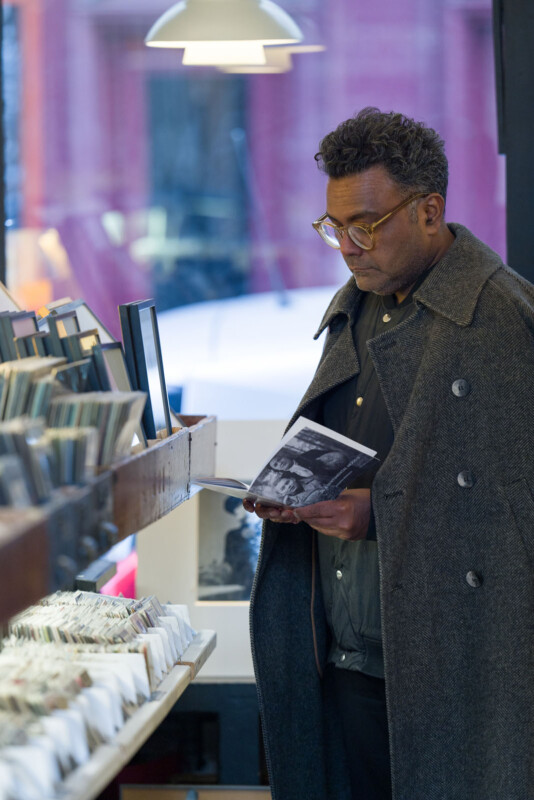 A man wearing glasses and a gray coat browses through records or CDs in a music store, holding one item and looking at its cover while standing by a display shelf.