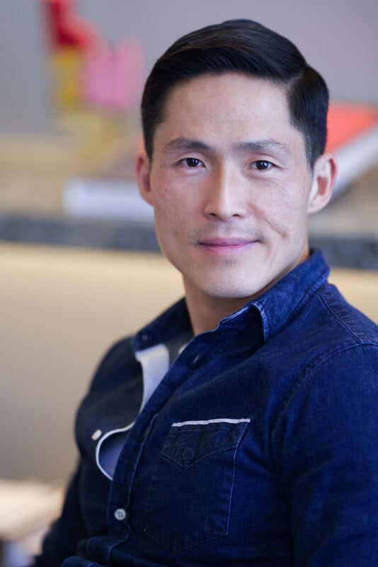 A man with short black hair wearing a dark blue denim shirt sits indoors, looking directly at the camera with a slight smile. The background is softly blurred with hints of colorful objects.